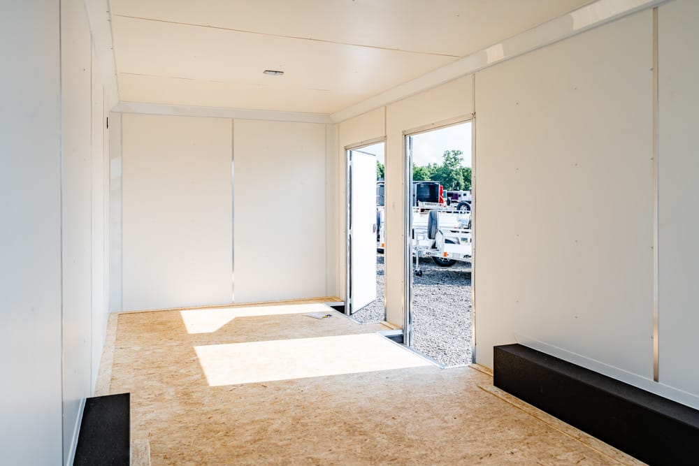 Interior of an empty white trailer with open doors, exposing a gravel lot and parked trucks outside.