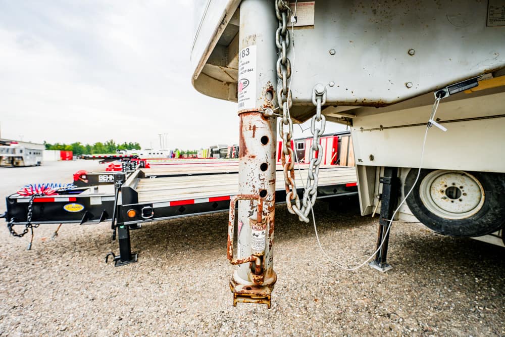 Rusted trailer hitch and chains on a parked flatbed trailer in a truck yard, with other equipment in the background.