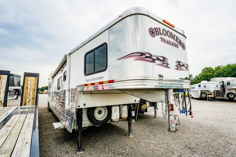 White Bloomer Trailers fifth-wheel RV with red flame graphics, parked on a gravel lot with other trailers in the background, hitch and stabilizers visible.”