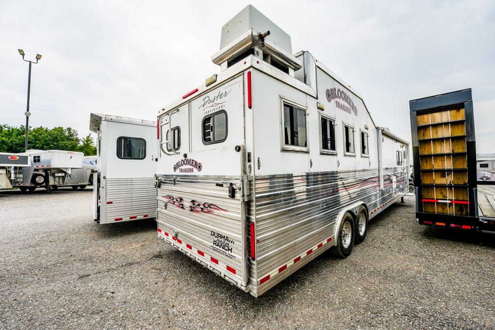 Silver, double-axle horse trailer with chrome siding and red accents parked on a gravel lot beside other trailers.
