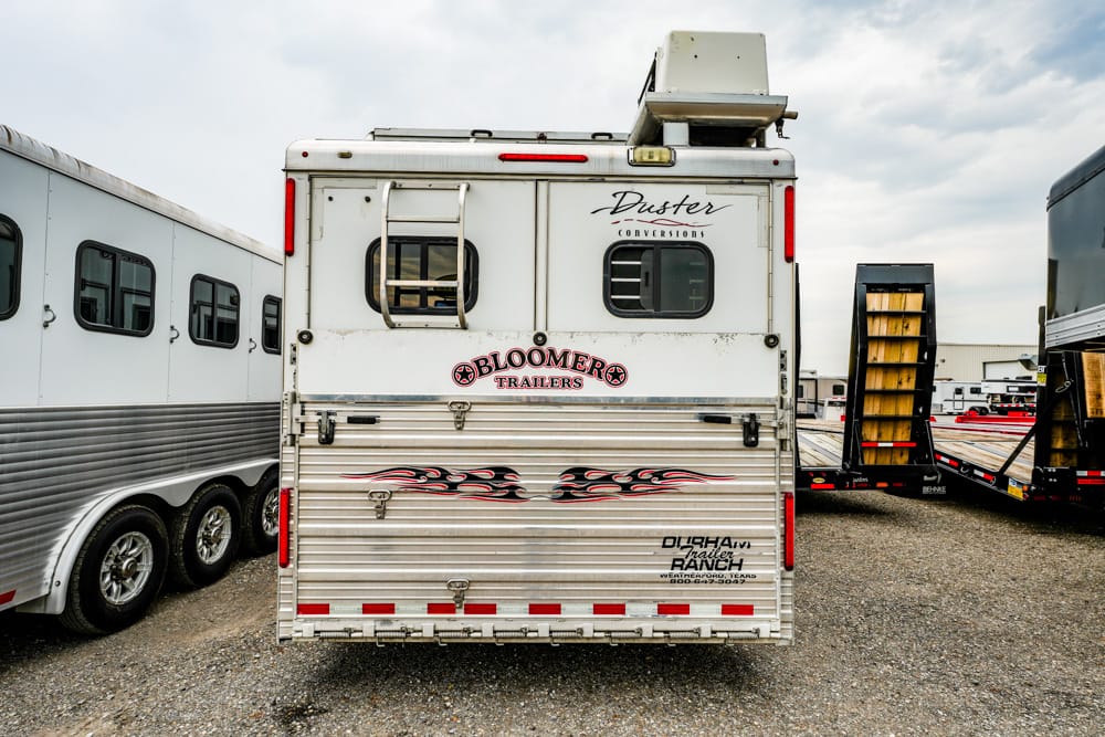 Rear view of a white horse trailer with chrome accents, flame decals, and 'Bloomers Trailers' logo, parked among other trailers.