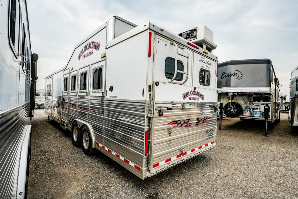 Rear view of a silver Bloomer horse trailer with flame decals parked in a gravel lot, surrounded by other RV trailers.