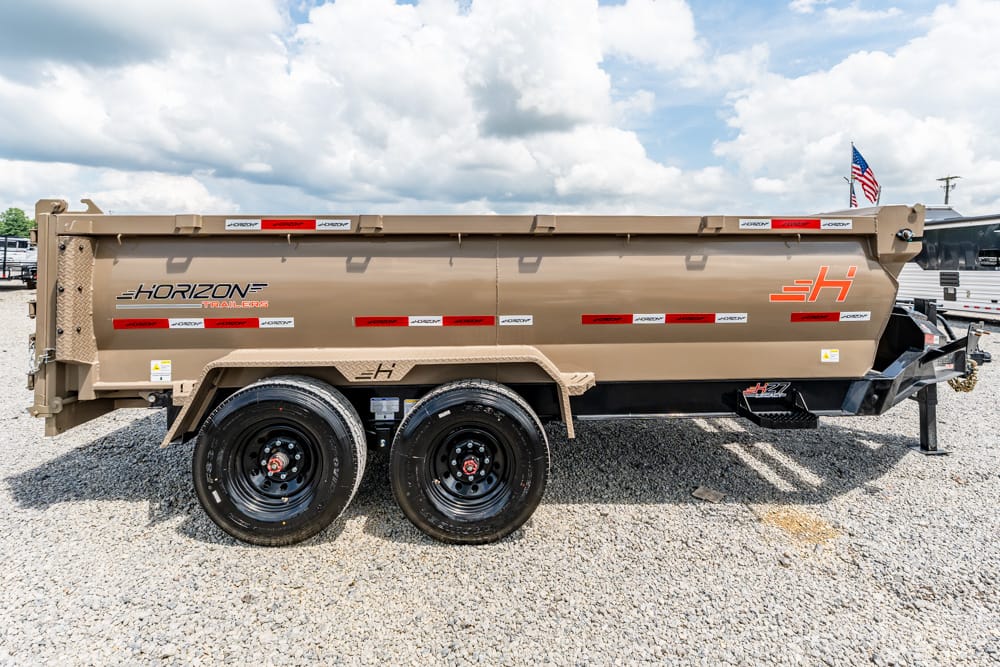 Tan Horizon dump trailer with dual axles and black wheels, parked on gravel with an American flag nearby in bright daylight