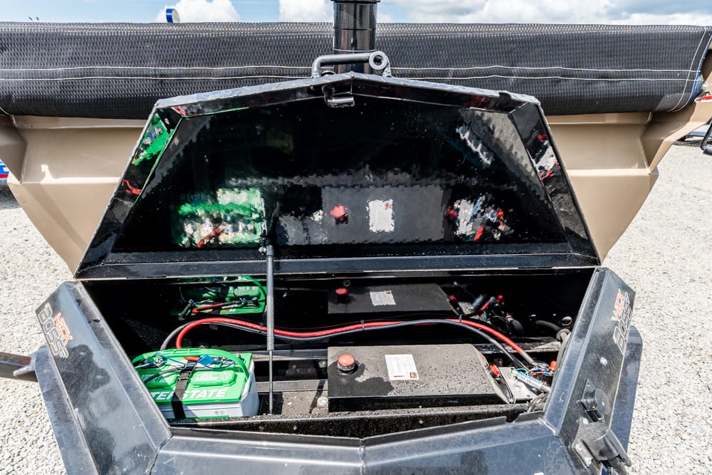Open battery compartment of a trailer, showing a green Interstate battery, red and black cables, and a dark storage area with a hitch nearby.