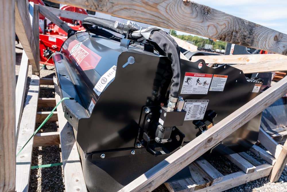Black metal industrial machine component with warning labels and hydraulic connections strapped in a wooden crate outdoors on gravel.