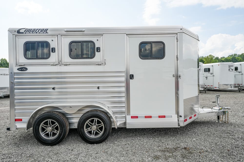 Two-axle aluminum horse trailer parked on gravel, showing side with two front windows and a side door, plus a hitch at the front.