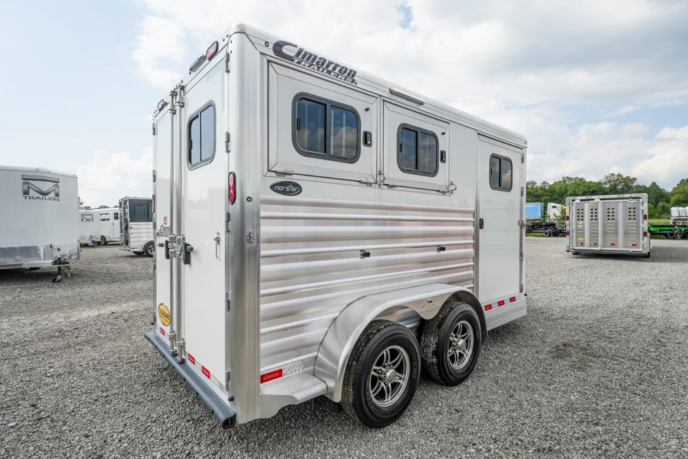 Rear three-quarter view of a silver aluminum two-axle horse trailer with small windows, parked on a gravel lot with other trailers in the background