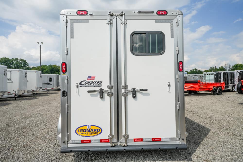 Rear view of a white enclosed cargo trailer with double doors, small window, and metal latches, on a gravel lot with other trailers nearby (logos visible).