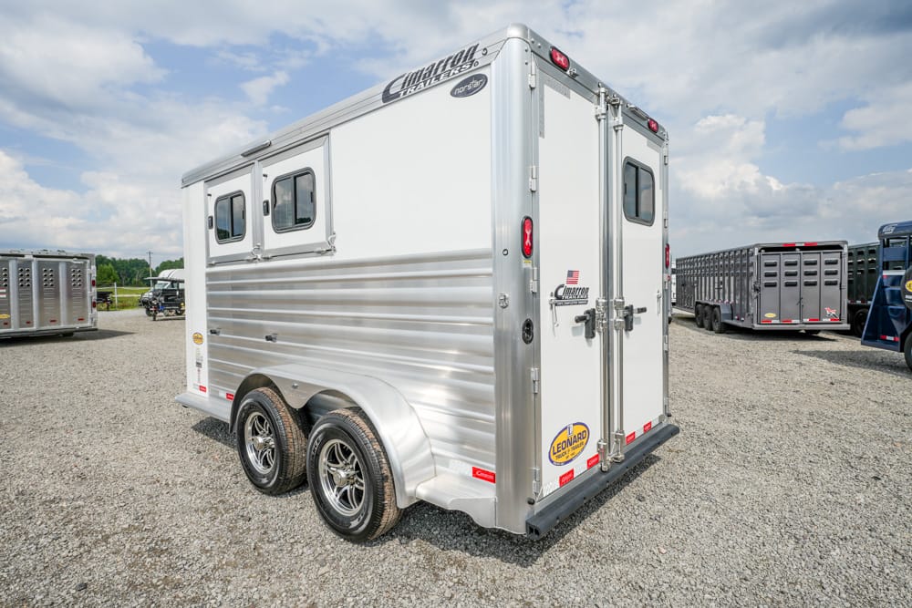 Silver dual-axle cargo trailer with tie-down doors, parked on gravel in a trailer lot under a cloudy sky