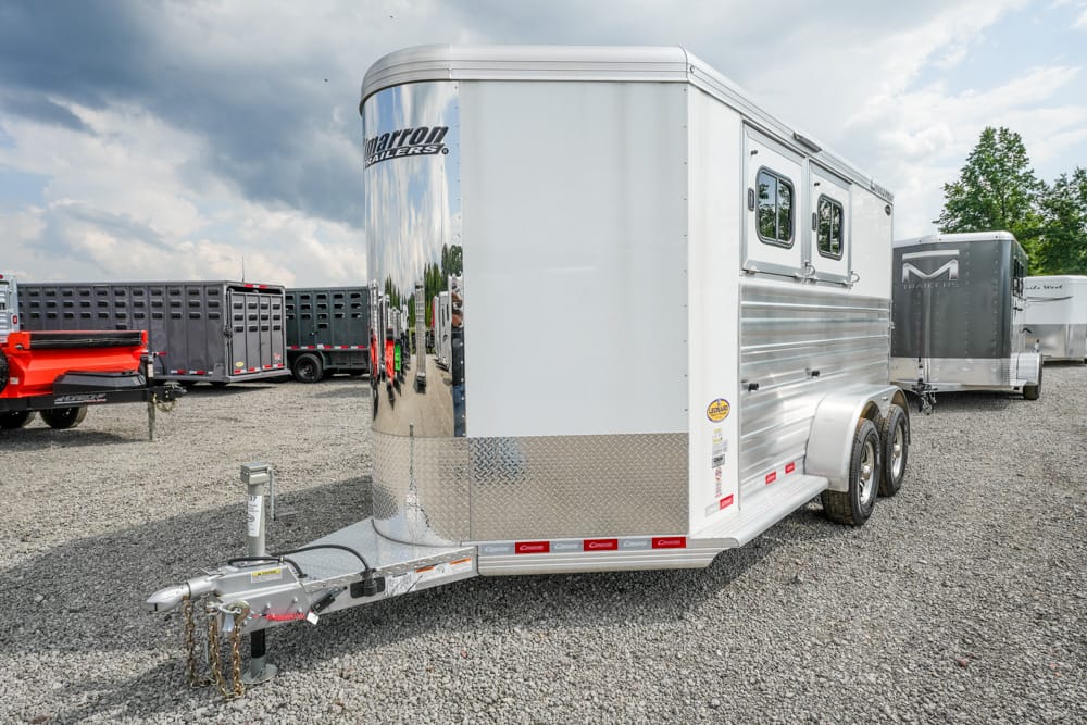 Silver dual-axle horse trailer in a gravel lot, front hitch and tongue visible in foreground.