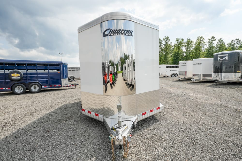 Front of a shiny Cimarron trailer with a diamond-plate lower panel, hitch in the foreground, in a gravel trailer lot with other trailers behind.