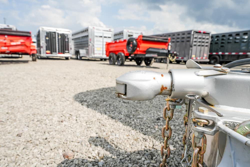 Close-up of a rusted trailer hitch with chains in the foreground; a gravel lot and several trailers parked in the background.