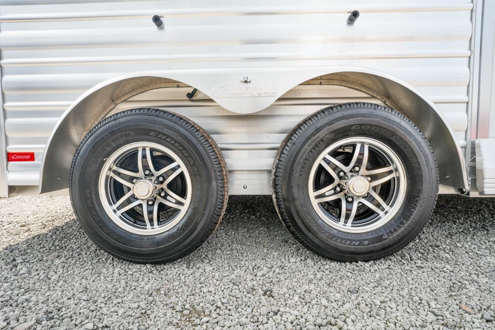 Twin trailer wheels with black tires and silver 5-spoke rims under a silver corrugated trailer, on gravel ground, with wheel fenders intact.