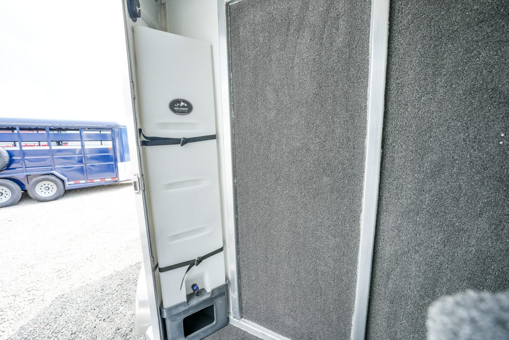 Open trailer door showing the inside wall with two strap locks and a small storage opening at the bottom; a blue trailer is visible outside.