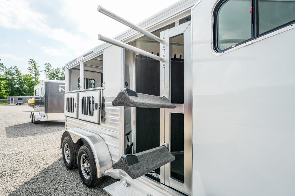 Silver horse trailer with open side stall door and fold-out steps on a gravel lot.