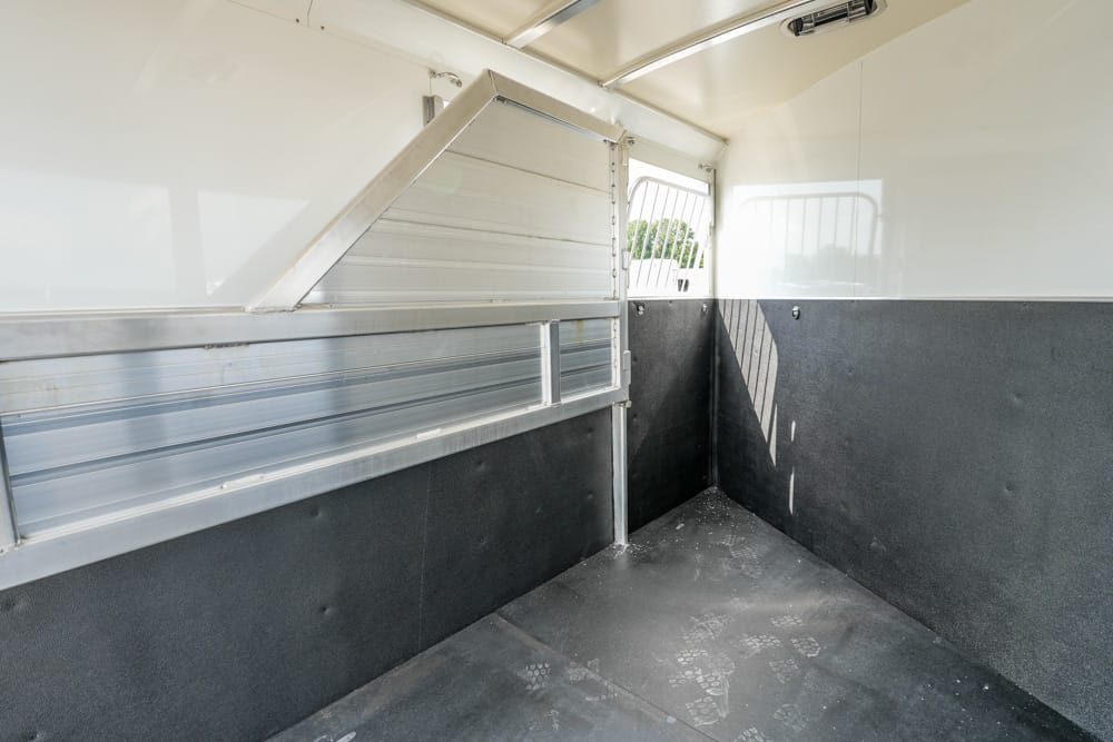 Interior of an empty horse trailer with a metal partition, white upper walls, and black rubber flooring near the corner.