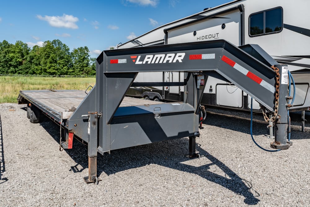 Gray Lamar trailer tongue and flatbed trailer on a gravel lot, with a white camper/RV in the background.