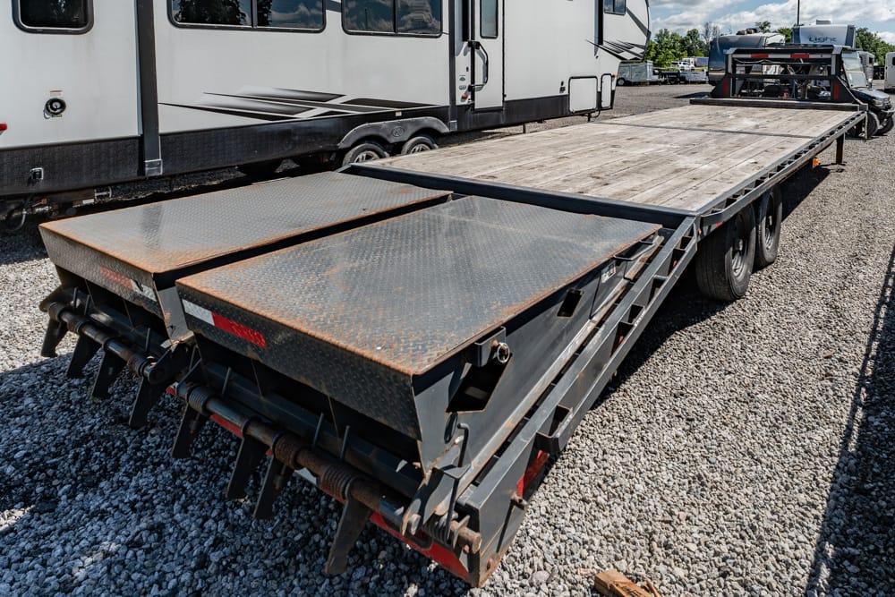 Steel flatbed trailer with rusted diamond-plate ramps, parked on gravel beside an RV.