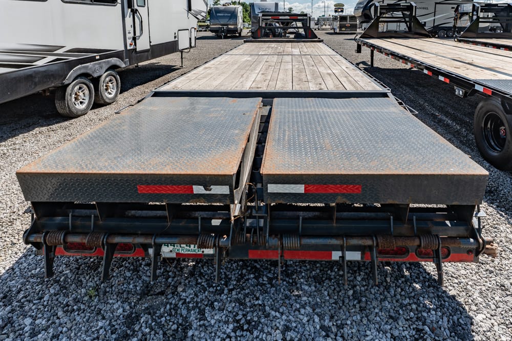 Rear view of a double-ramp, diamond-plate trailer bed with rust, parked on a gravel lot among other trailers (vehicle loading ramps).