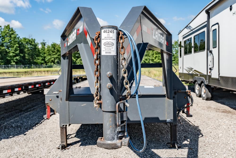 Heavy equipment trailer hitch with rusted chains and a blue cable, tag 46245, in a gravel lot with a nearby RV trailer.