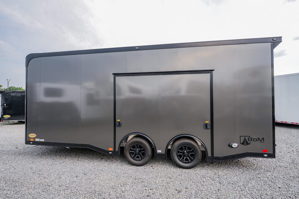 Gray enclosed cargo trailer with two axles and a side door on a gravel lot, branded ROM near the back.
