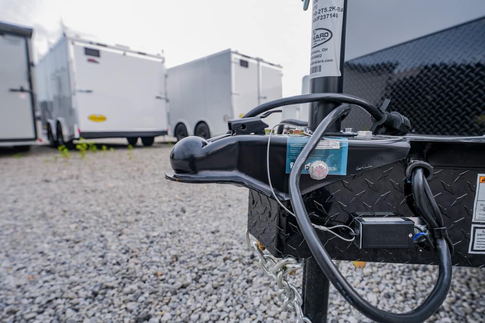 Close-up of a trailer hitch with wiring on a diamond-plate trailer in a gravel lot, white trailers in the background.