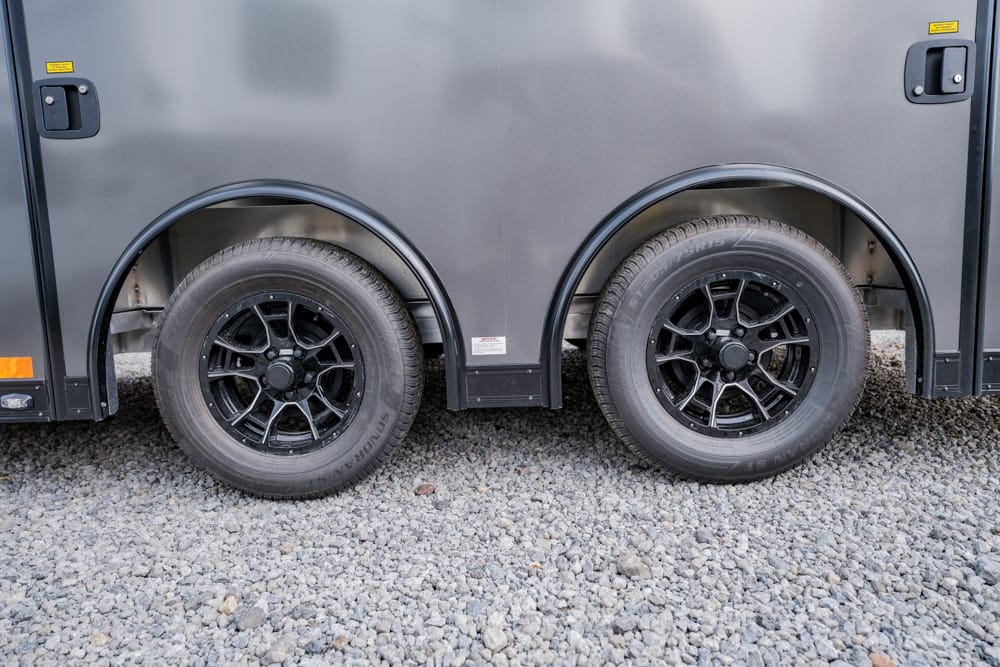 Dual axles with black alloy wheels on a grey trailer, parked on gravel surface