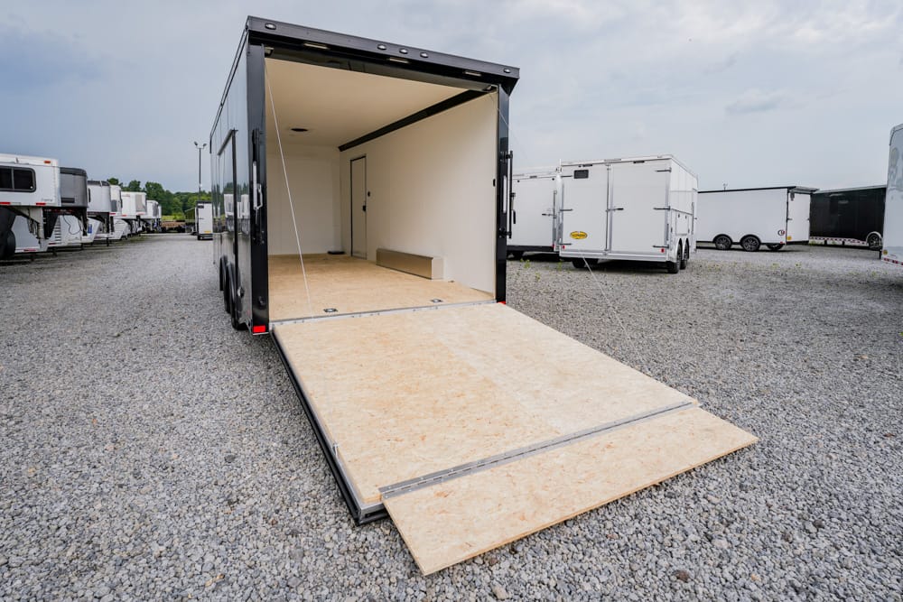 Open black cargo trailer with ramp down, empty interior, on a gravel lot among other trailers.
