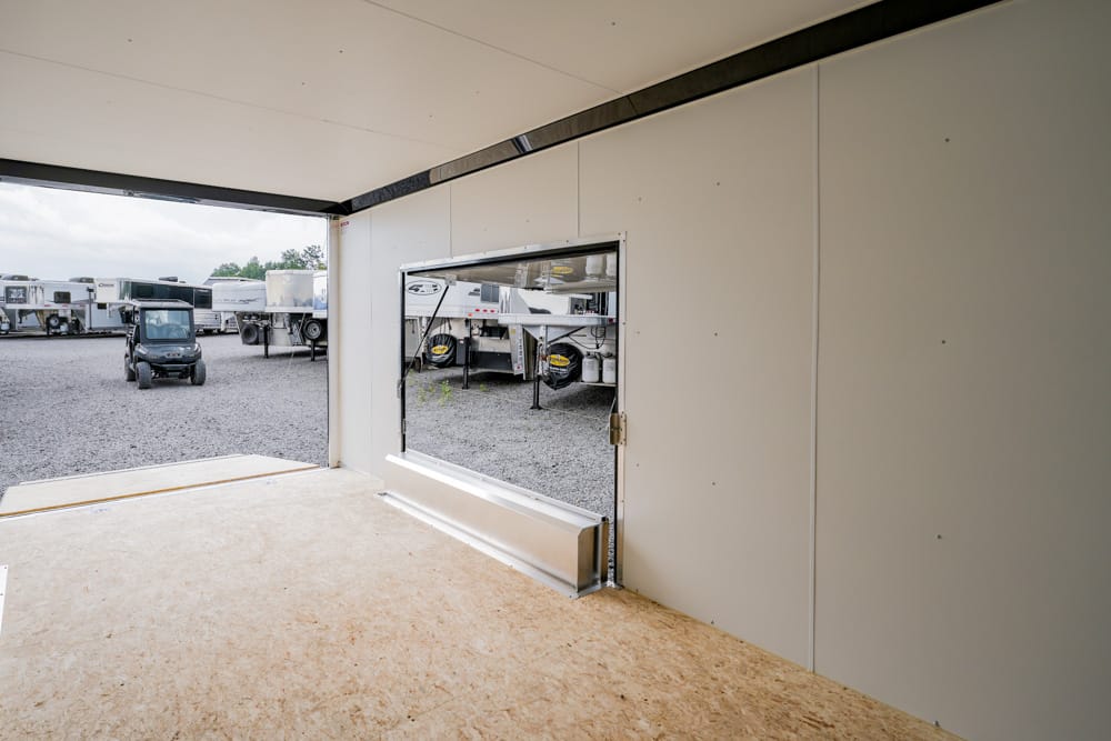 Interior of a beige, paneled space with an open wall showing a gravel yard full of horse trailers and a small utility cart nearby