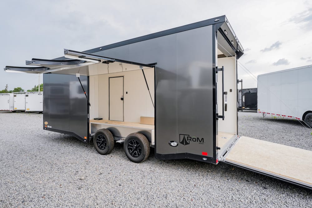 Gray, two-axle enclosed trailer with ramp lowered, interior bench and open back door wi th a gravel lot