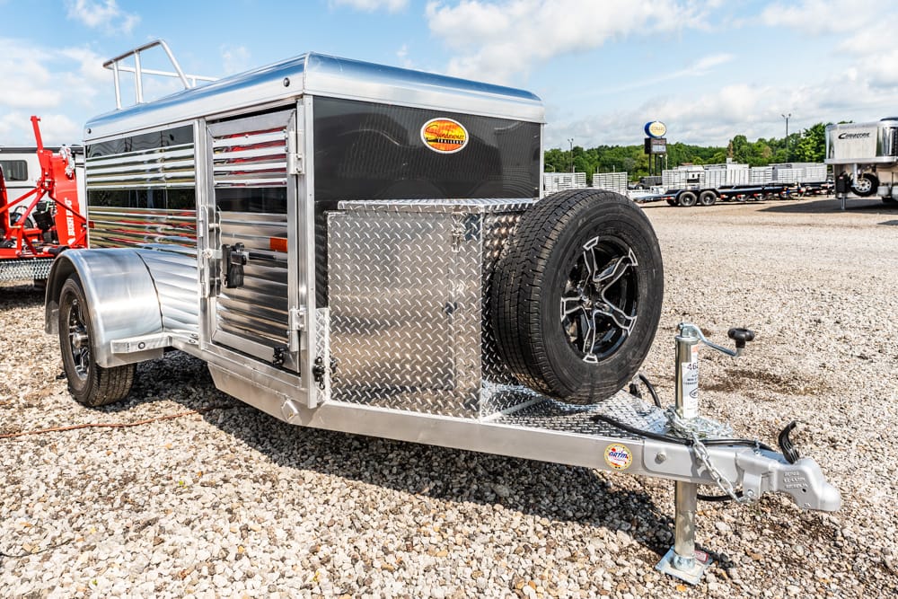 Aluminum utility trailer with a spare tire mounted on the front and a diamond‑plate toolbox, parked on a gravel lot by other trailers in the background.