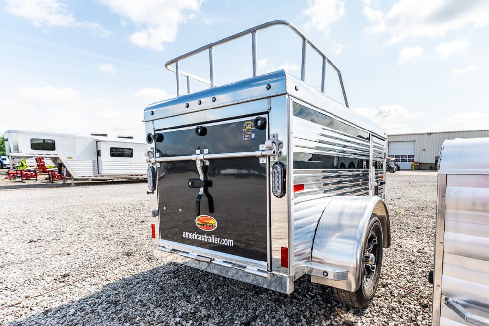 Rear view of a shiny aluminum utility trailer with a top railing, parked on a gravel lot; back doors closed and americatrailer.com sticker visible.