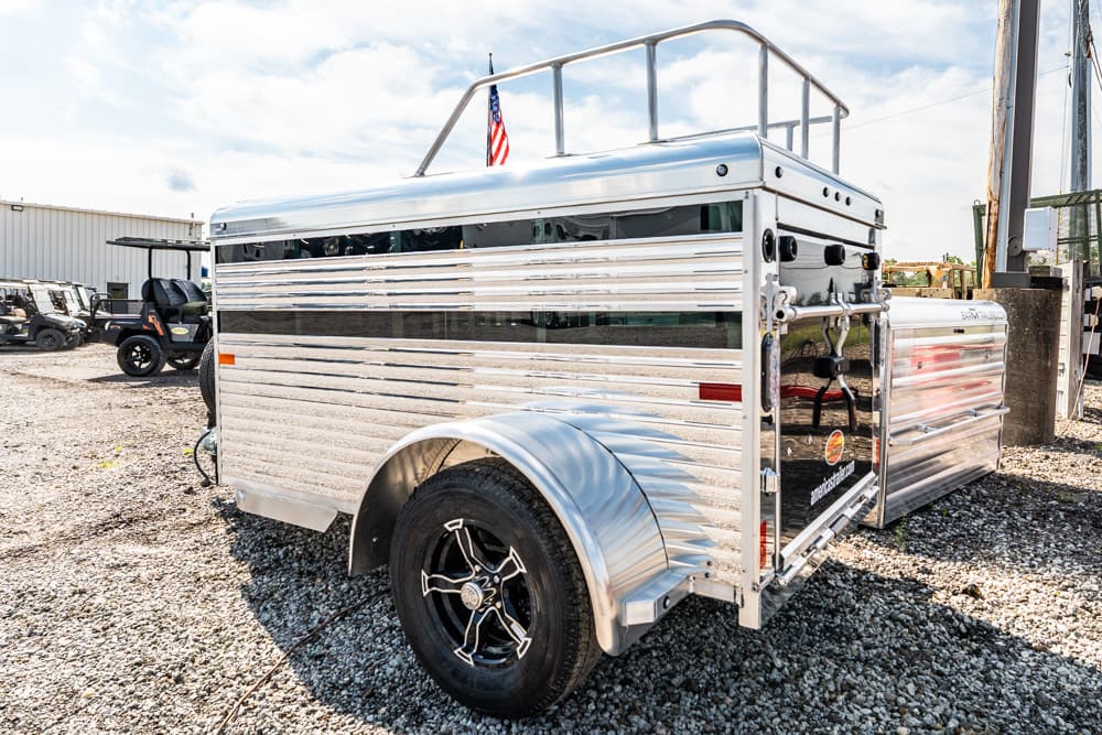 Shiny aluminum livestock trailer with rounded top rails parked on a gravel lot, American flag on top and other vehicles in the background.