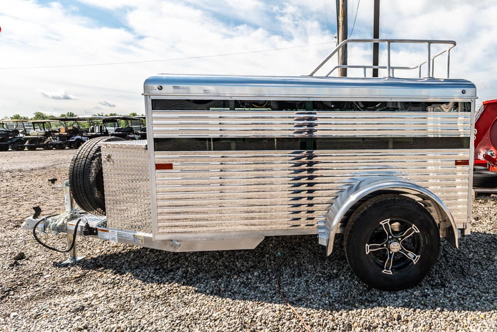 Shiny aluminum enclosed trailer on a gravel lot, with a spare tire mounted at the front and a towing hitch