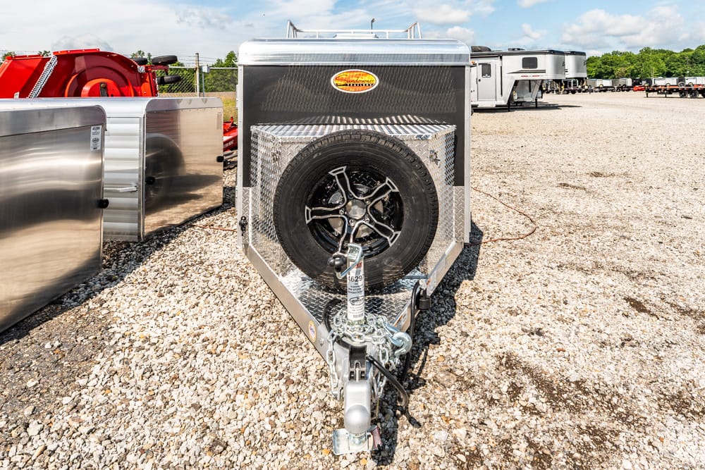 Front view of a silver trailer with a mounted spare tire on diamond-plate front, hitch and propane tank in a gravel lot.