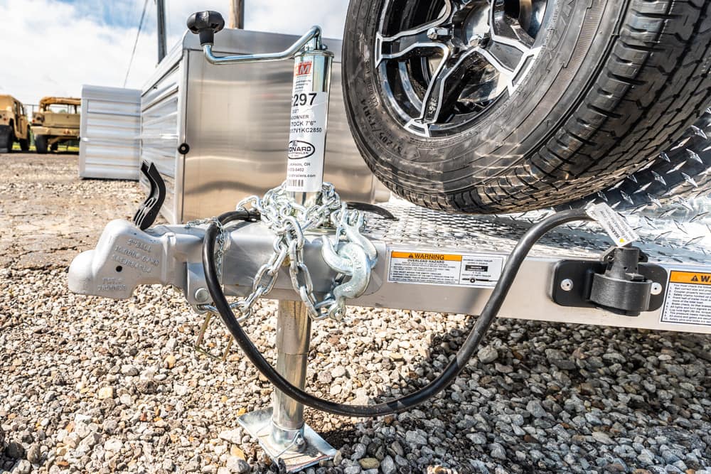 Trailer hitch and jack on a utility trailer with a spare tire mounted on the diamond-plate deck; safety chains are wrapped around the coupler on a gravel lot.