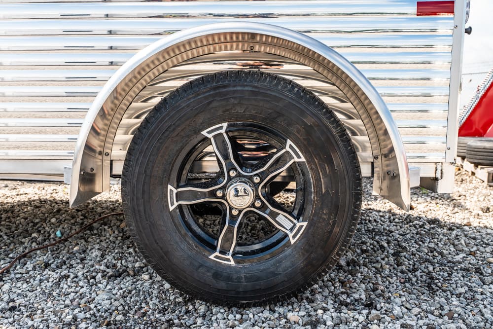 Close-up of a trailer wheel: black tire on a silver five-spoke alloy rim under a chrome fender on gravel ground.