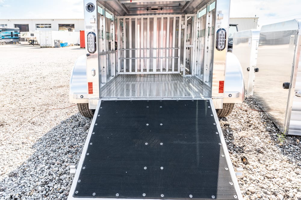 Silver horse trailer with the rear ramp lowered, showing empty stalls inside and a non-slip loading ramp on a gravel lot