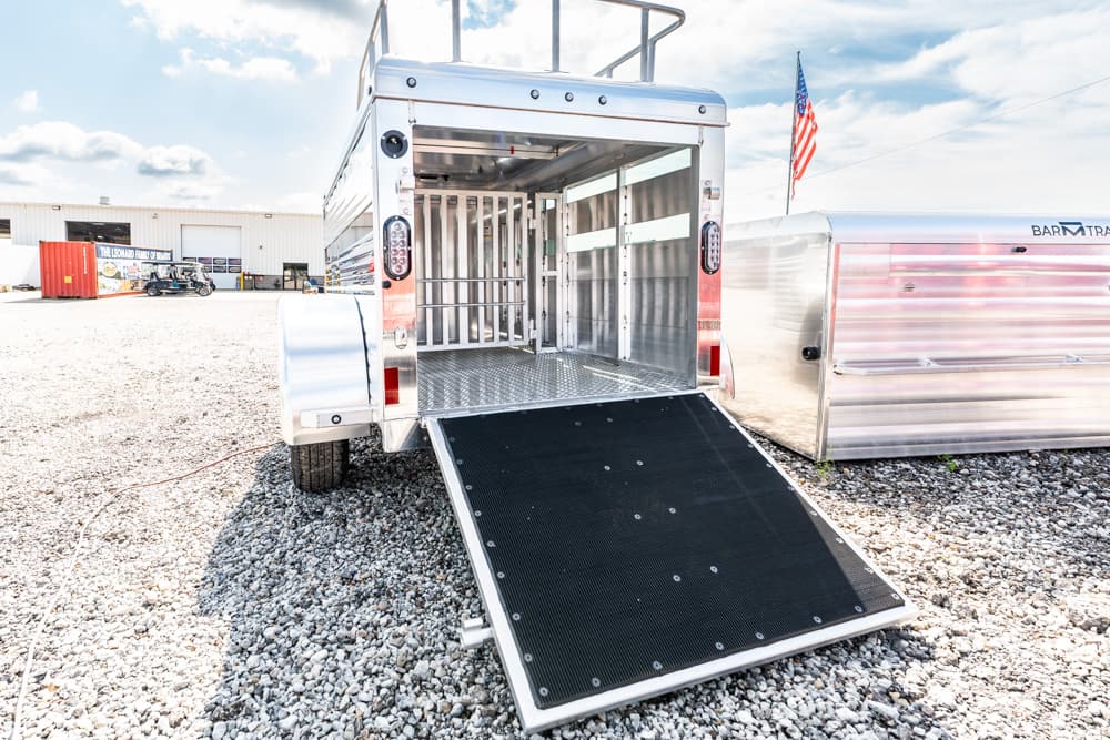 Open horse trailer with ramp down, showing empty stalls on a gravel lot with another trailer nearby and an American flag in the background.