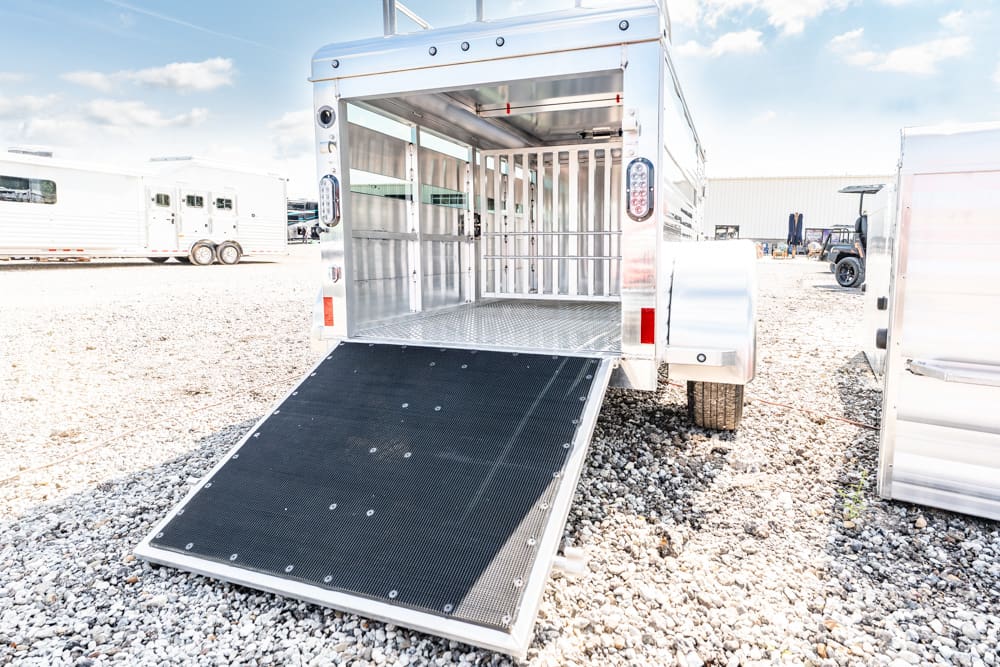 Aluminum horse trailer with its rear ramp lowered, showing empty interior stalls on a gravel lot.