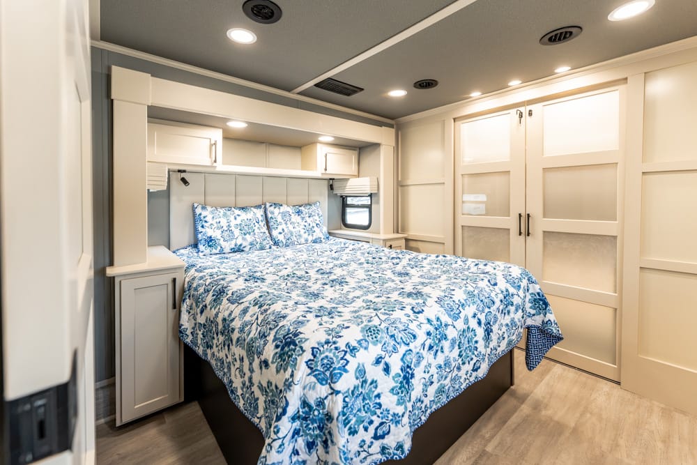 RV bedroom with a queen bed, white cabinetry, and blue floral bedding; built-in nightstands and overhead storage create a cozy, organized space.