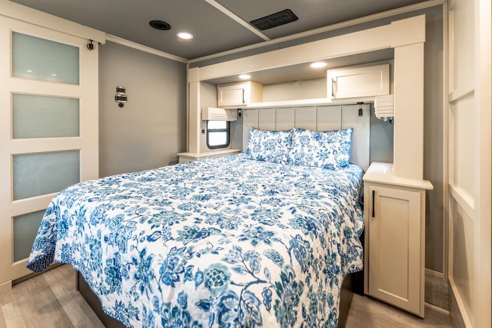 Queen bed with blue floral duvet in a compact RV bedroom with built-in white storage and windows above.
