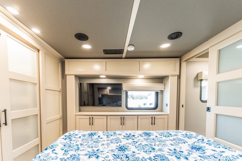RV bedroom interior with a cream built-in cabinet wall, window opposite the bed, and a blue floral bedspread at the foot of the bed. Lush lighting overhead.