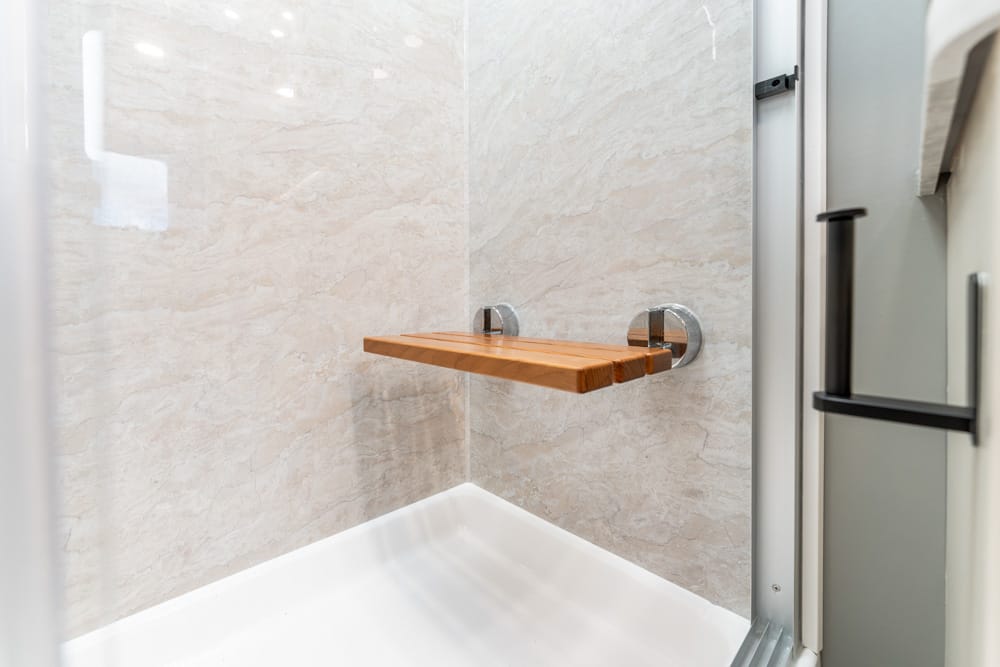 Wooden folding shower seat mounted to tiled shower wall inside a glass-enclosed shower stall