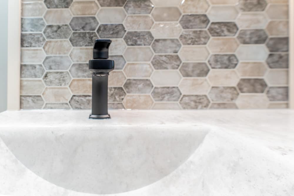 Black matte bathroom faucet on a white sink with a beige-gray hex tile backsplash behind it.