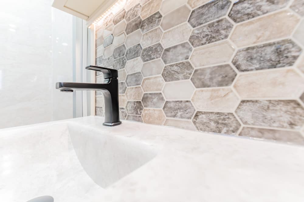Black modern faucet over a light stone sink with a hexagonal tile backsplash in neutral tones.