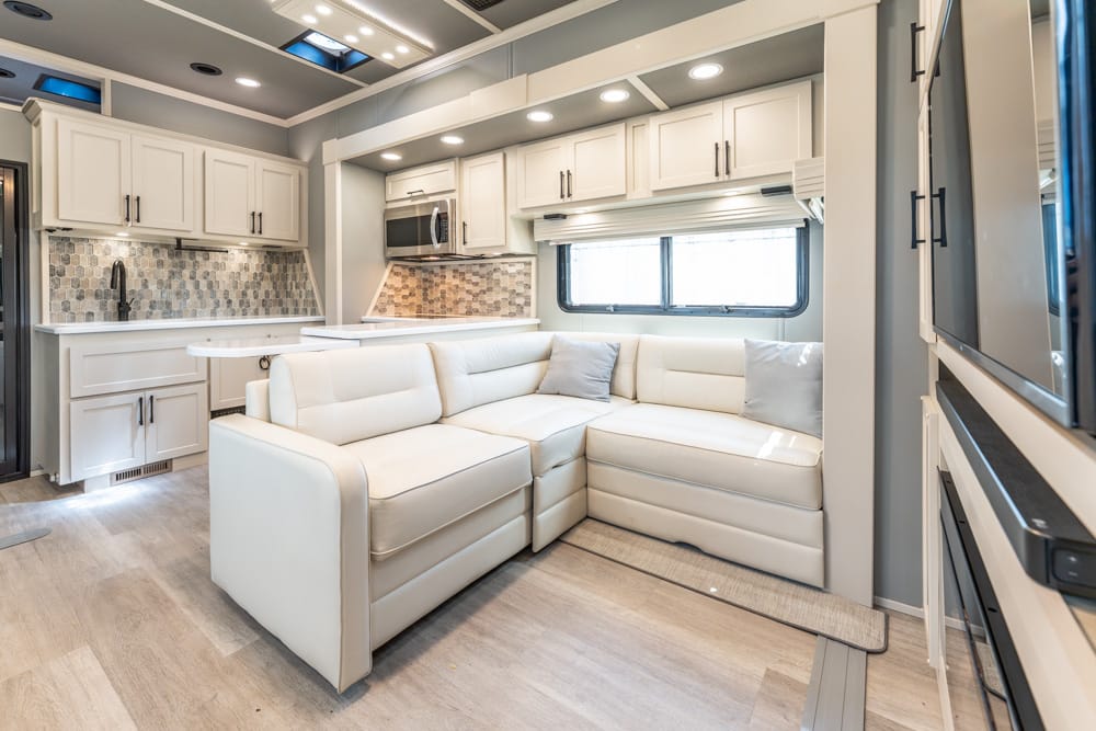 Bright RV living area with a white leather sectional, compact kitchen, and tiled backsplash undercabinet lighting