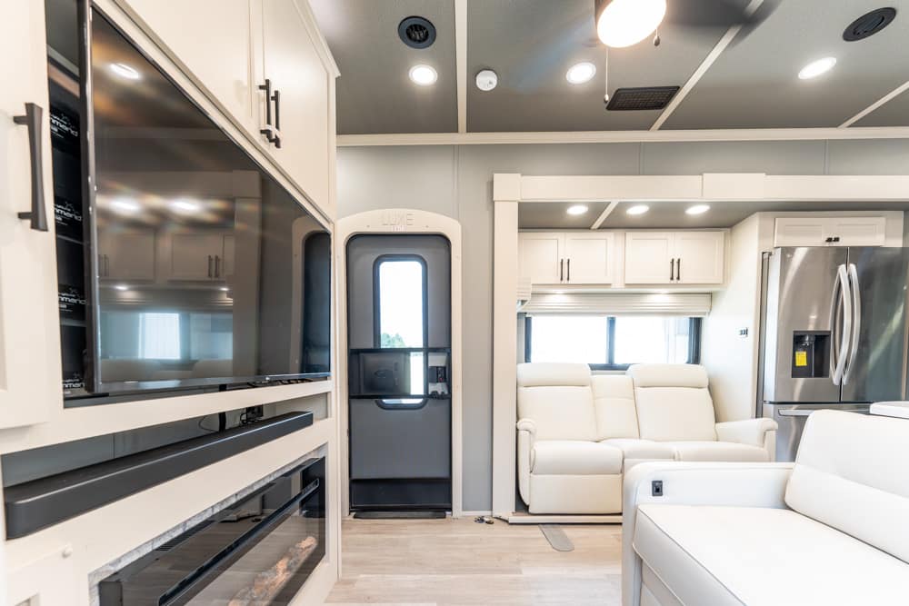 Bright, modern RV interior with white leather sofas, a large wall-mounted TV, and a stainless fridge.
