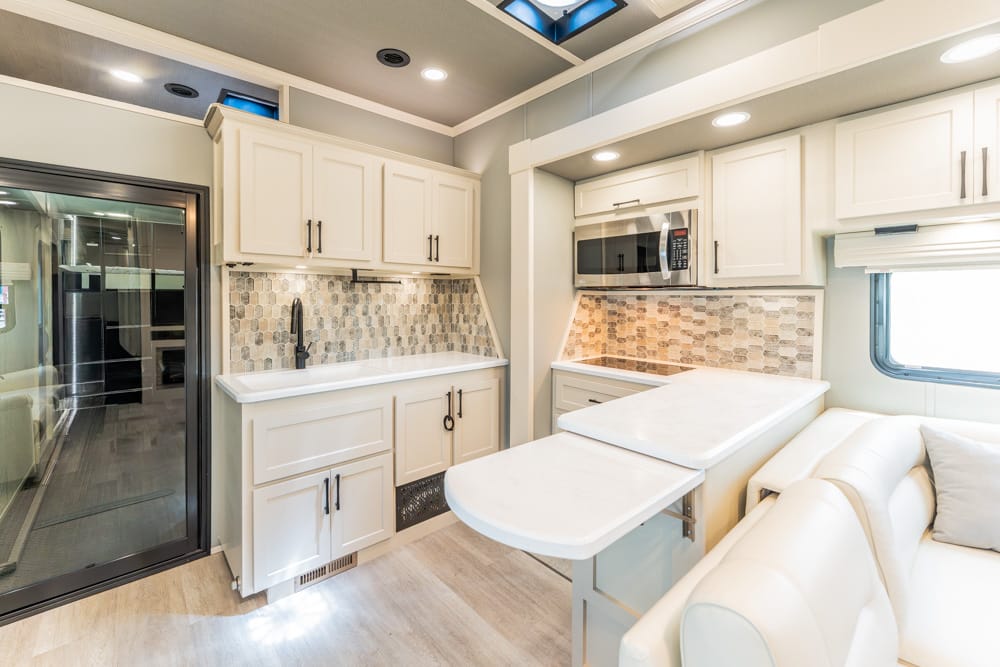 Modern RV kitchen with white cabinets, tile backsplash, and a fold-out island beside a cream sofa.