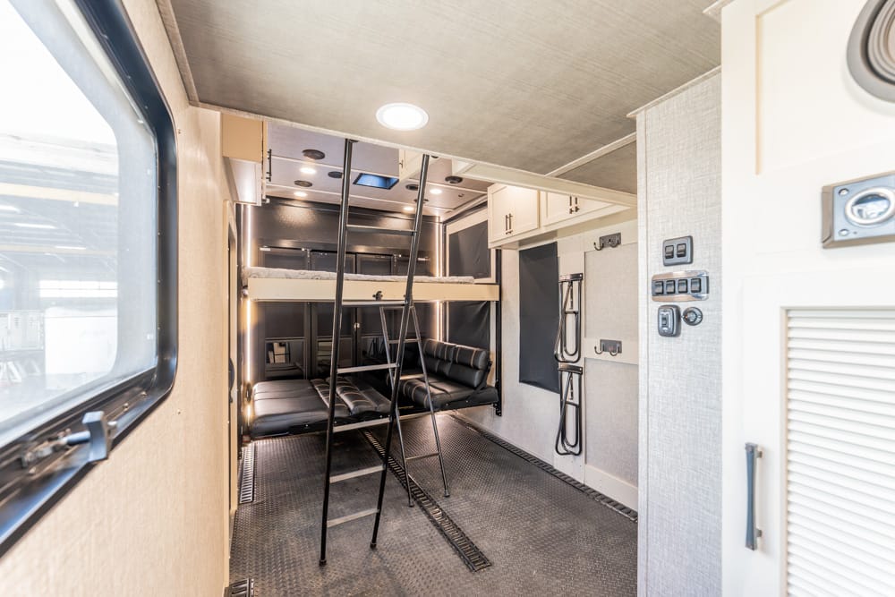 Interior of a modern camper trailer with a loft-style bunk bed, metal ladder, and black leather seating area.